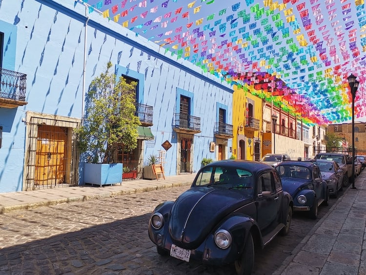 A colourful street in Oaxaca with multicoloured bunting and a row of beetle cars parked on the road