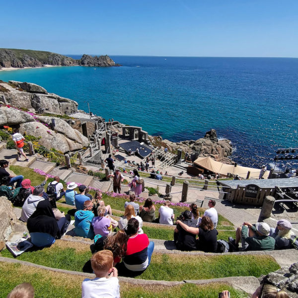 Looking down over the Minack Theatre, Cornwall. People are sat on grass growing over the concrete seats, looking over the theatre and the bright blue sea behind