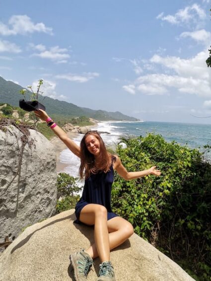 Giselle sat on a large rock waving her cap out in front of a wild sea and coastline view in Tayrona Natural National Park